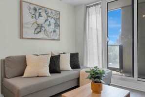 A living room with a grey sofa, a painting on the wall, a window with curtains, and a plant on a table. - City Center – Walkable 2BR, 5 min to Eaton Centre (Toronto)
