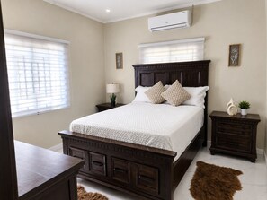A bedroom with a large bed, bedside tables, a nightstand, an air conditioning unit, and a window with blinds.