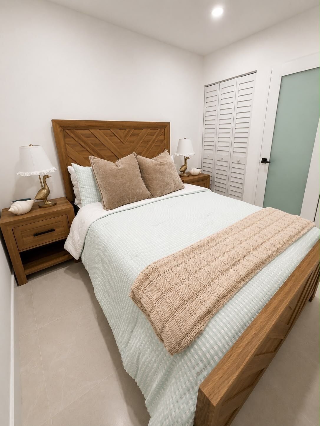 A bedroom with a wooden headboard, a bed with a light-colored quilt, two bedside tables with lamps, and a closet with sliding doors.