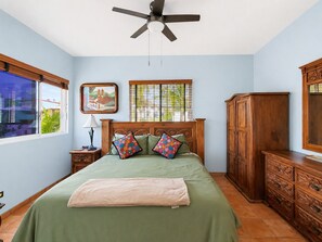 A bedroom with a bed, a ceiling fan, a nightstand, a lamp, a window with blinds, a wooden cabinet, and a framed picture on the wall. - Beautiful Apartment with amazing views ! A hidden gem! (San José del Cabo)