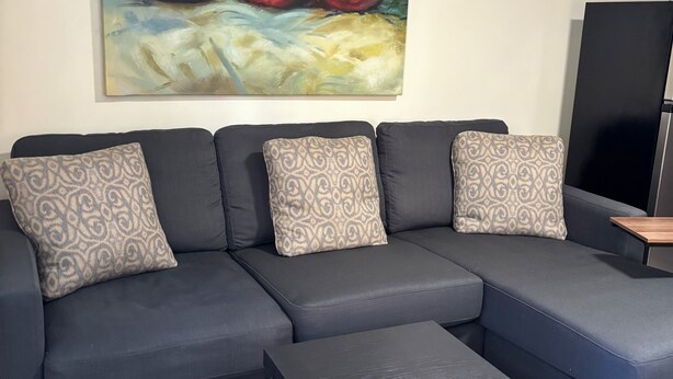 Ein modernes Wohnzimmer mit einem Ecksofa, gemusterten Kissen, einem Couchtisch und einem großen Gemälde mit Früchten an der Wand.