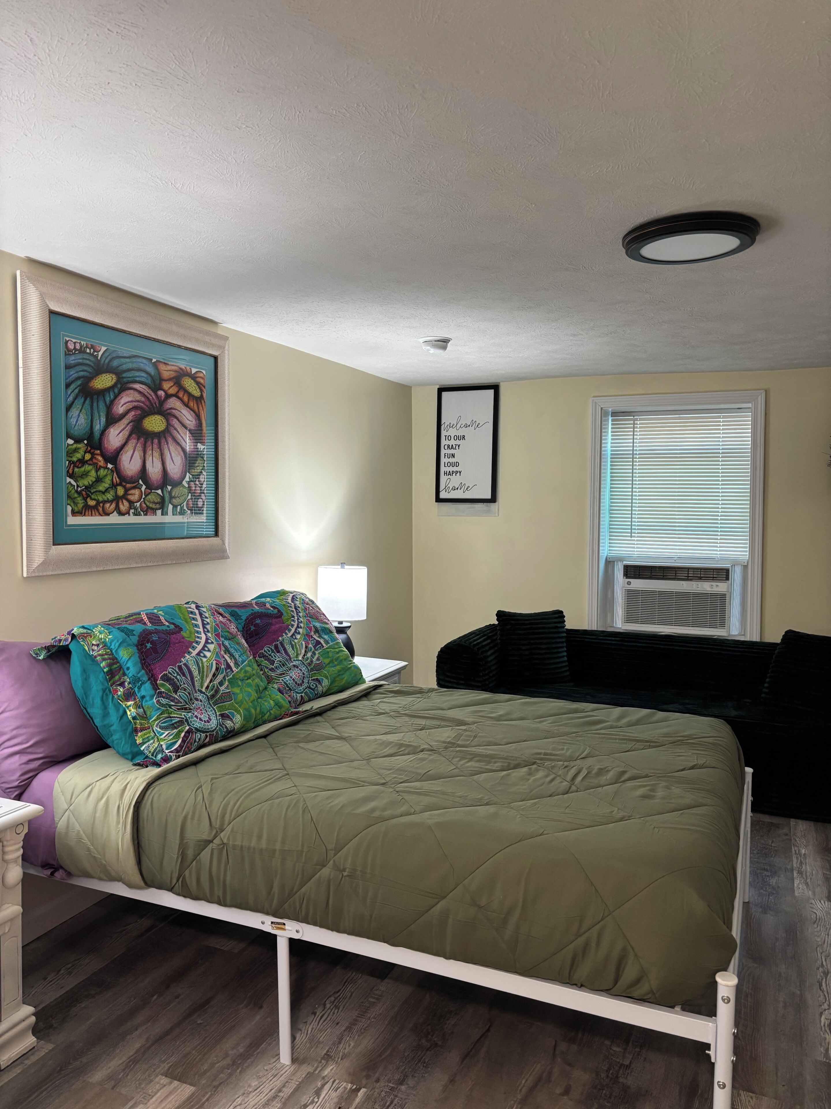 A bedroom with a bed, a nightstand, a wall-mounted lamp, a framed welcome sign, a window with blinds, and a ceiling light.