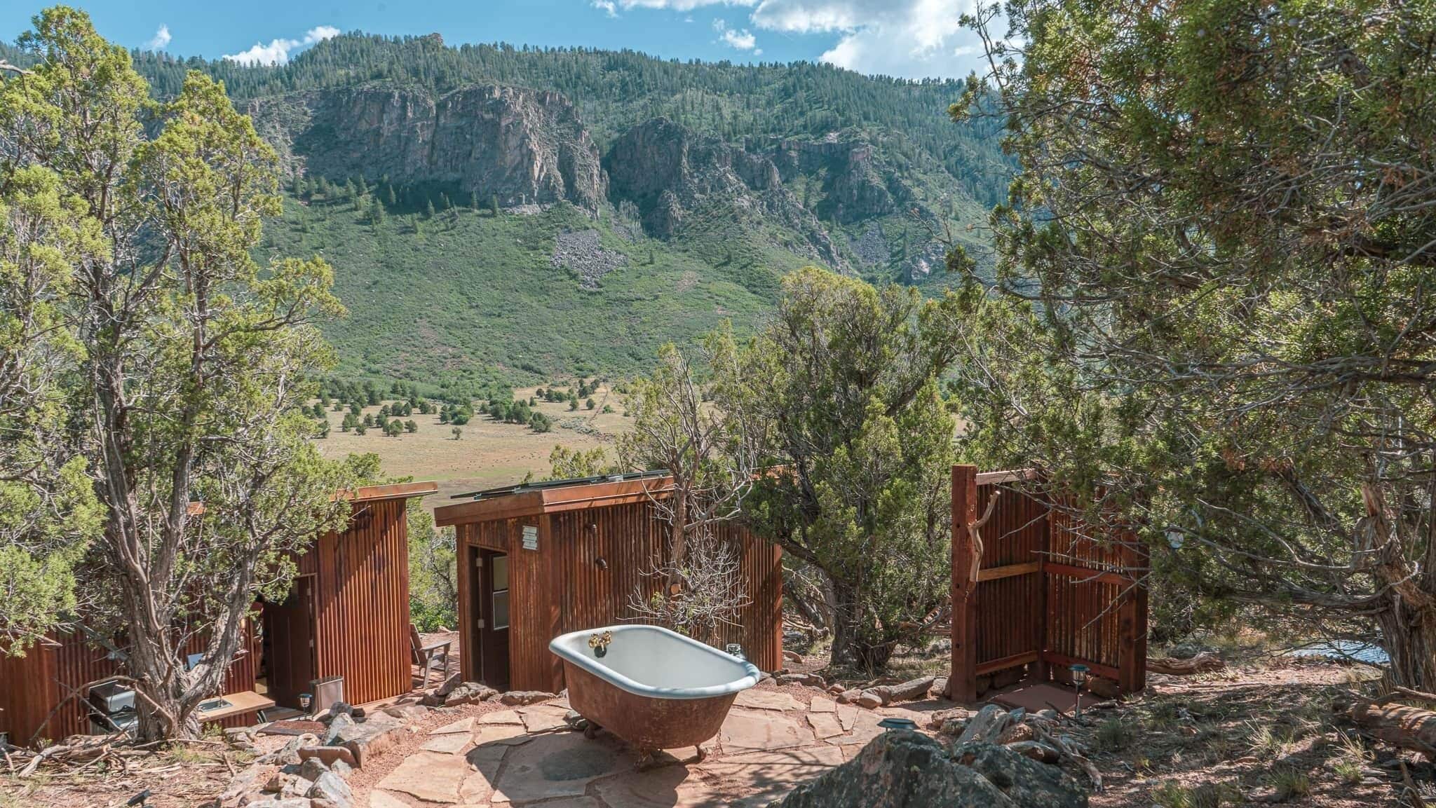 A rustic cabin with a stone patio, a bathtub, and wooden gates leading to a mountainous landscape.