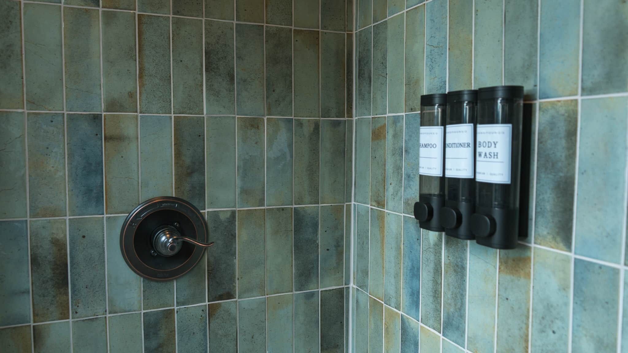 Un baño con pared de azulejos, desagüe de ducha y tres botellas etiquetadas como champú, acondicionador y gel de baño.