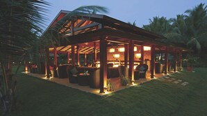 A covered outdoor seating area with tables and chairs, surrounded by palm trees. - Tropicana Resort and Spa Alibaug (Alibag)