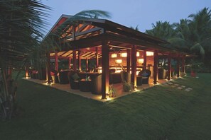 A covered outdoor seating area with tables and chairs, surrounded by palm trees. - Tropicana Resort and Spa Alibaug (Alibag)
