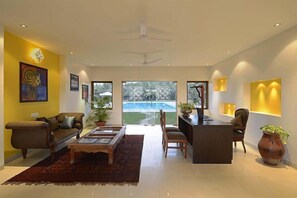 A modern living room with a sofa, coffee table, and a desk area. There is a view of a swimming pool through the glass doors. - Tropicana Resort and Spa Alibaug (Alibag)