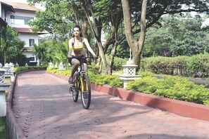 A person riding a bicycle on a paved pathway surrounded by greenery and a building in the background. - Tropicana Resort and Spa Alibaug (Alibag)