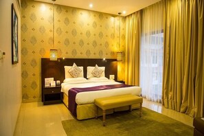 A hotel room with a large bed, a nightstand, a bench, and a window with curtains. - Tropicana Resort and Spa Alibaug (Alibag)