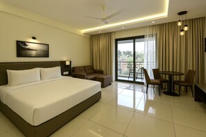 A hotel room with a large bed, a sofa, a dining table with chairs, a balcony with curtains, and a ceiling fan. - Tropicana Resort and Spa Alibaug (Alibag)