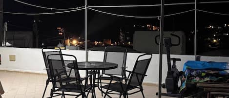 An outdoor seating area with a table and chairs, a covered bicycle, and a view of a cityscape at night.