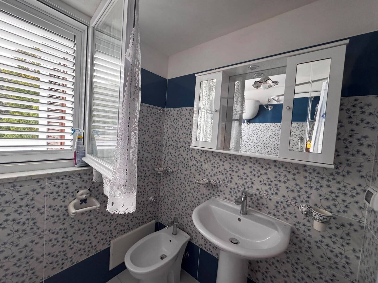 A bathroom with a bidet, a sink, a mirror, and a window with blinds.