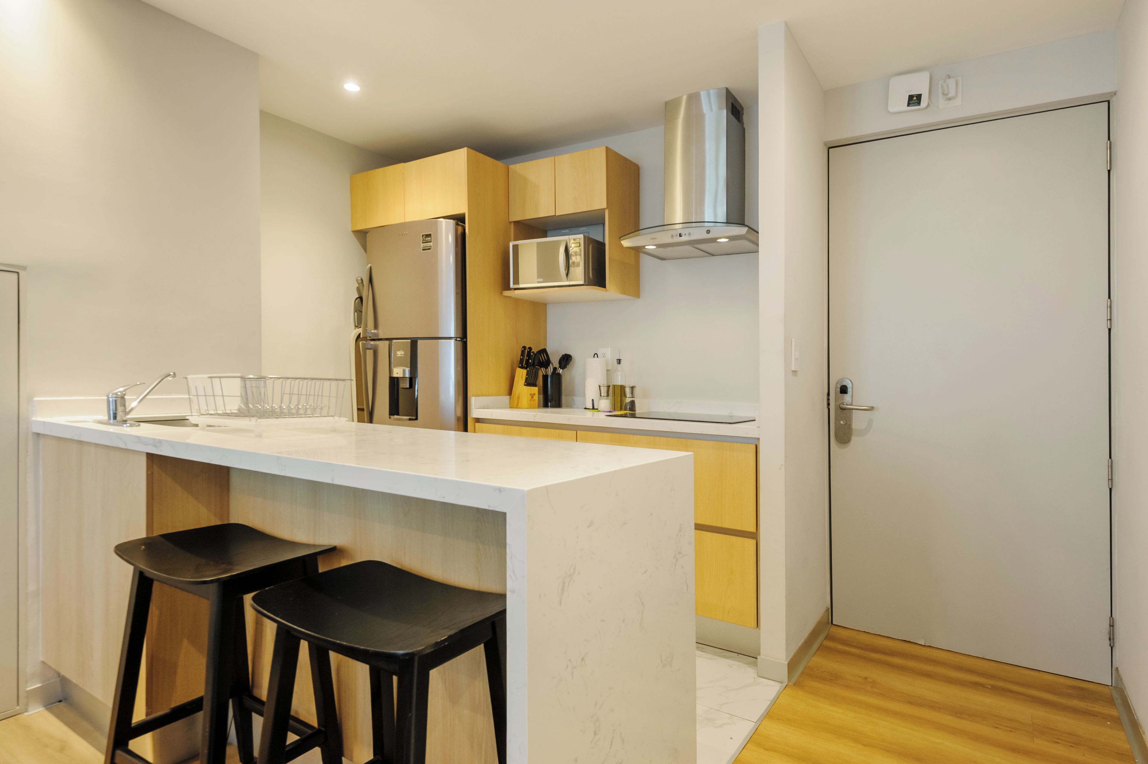 Una cocina compacta con una isla central, dos taburetes de bar, un microondas y una campana extractora de acero inoxidable.