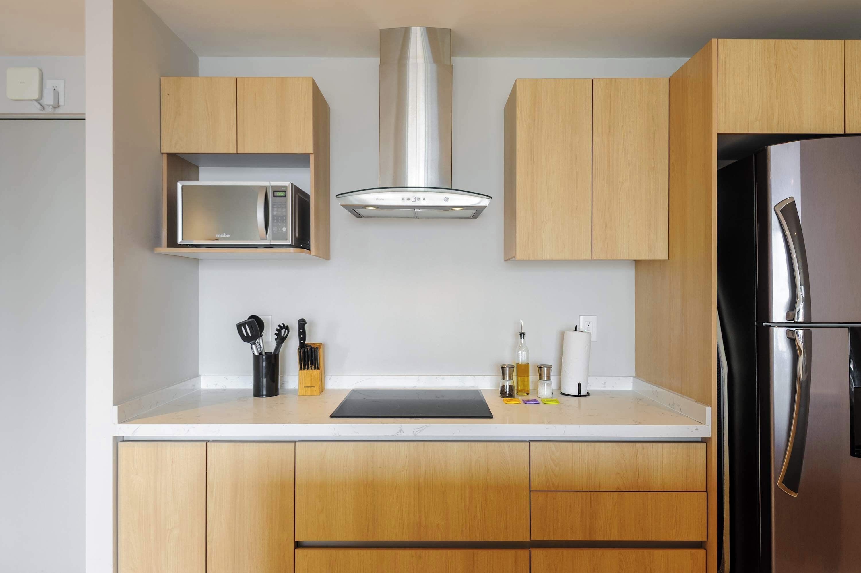 A modern kitchen with wooden cabinets, a stainless steel range hood, a microwave, a refrigerator, and a black cutting board.