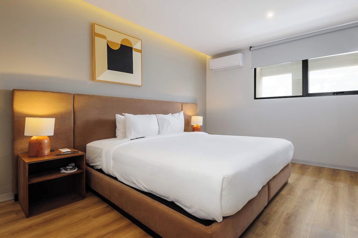 Quarto de hotel com uma cama grande, duas luminárias de cabeceira, uma mesinha de cabeceira, um quadro na parede e um ar condicionado.