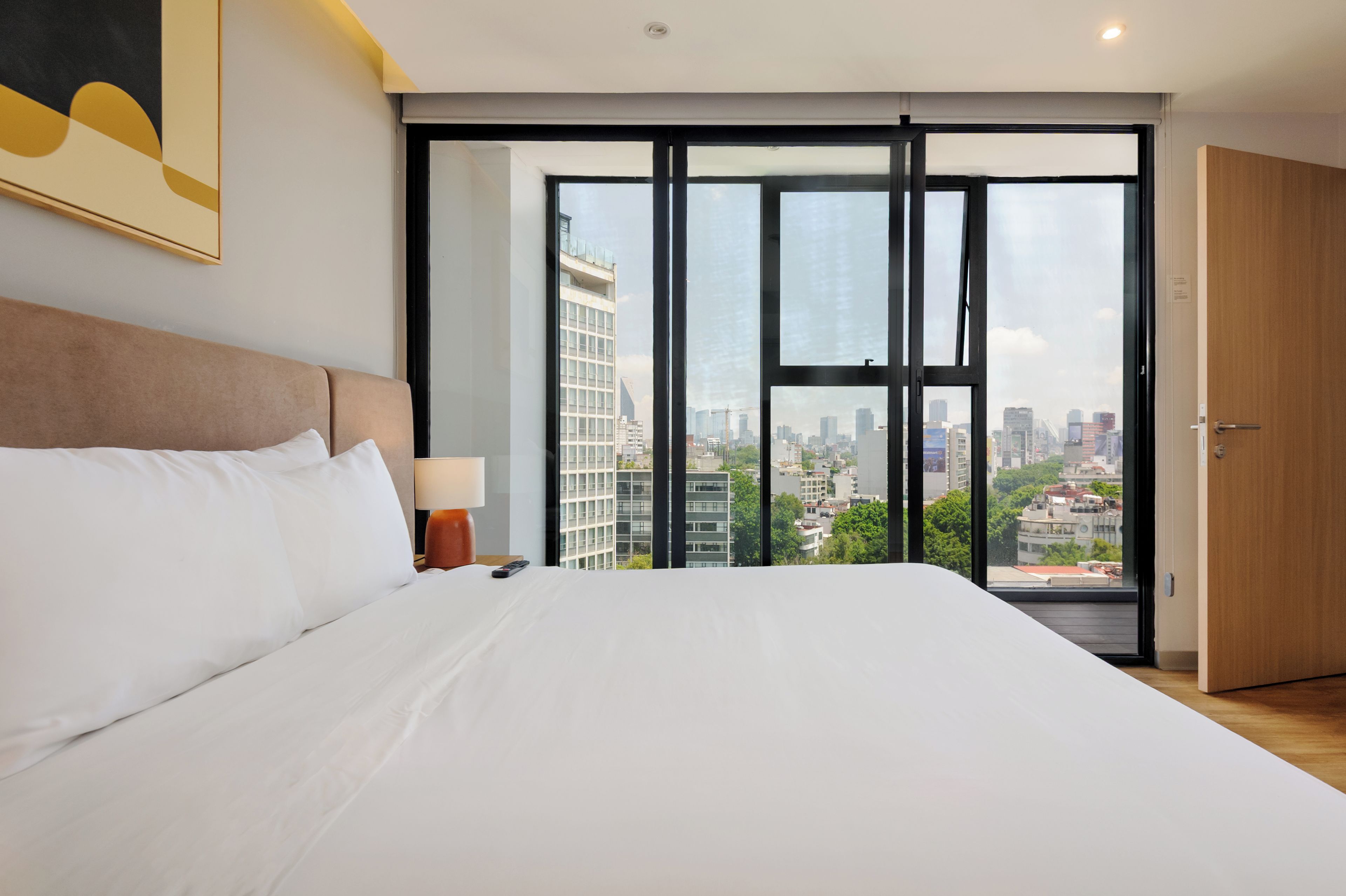Quarto de hotel com uma cama grande, mesa de cabeceira, abajur e vista da cidade através de janelas do chão ao teto.