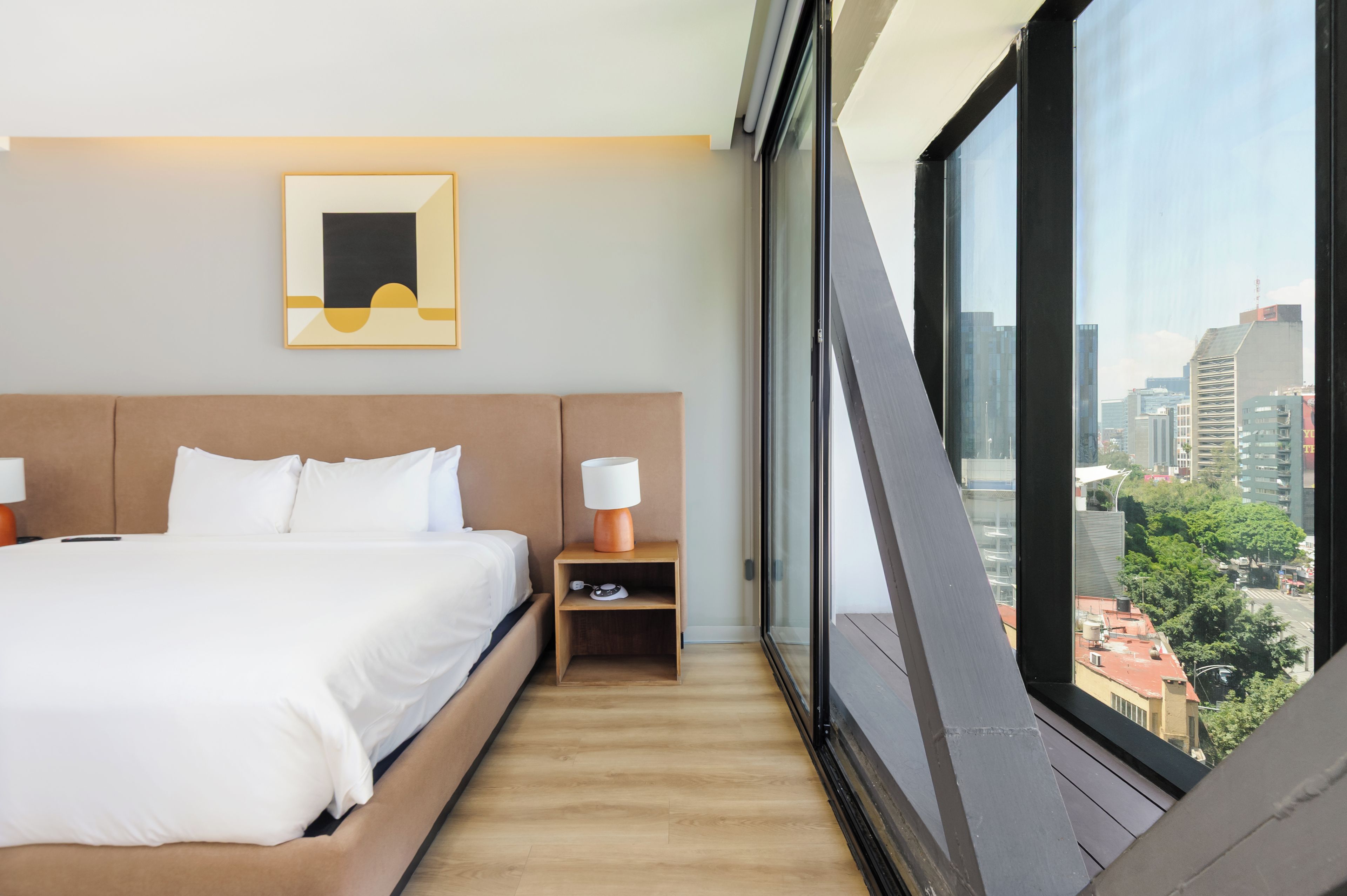 Ein modernes Hotelzimmer mit einem großen Bett, einem Nachttisch mit Lampe, einer kleinen Ablagefläche und Blick auf die Stadt durch ein großes Fenster.