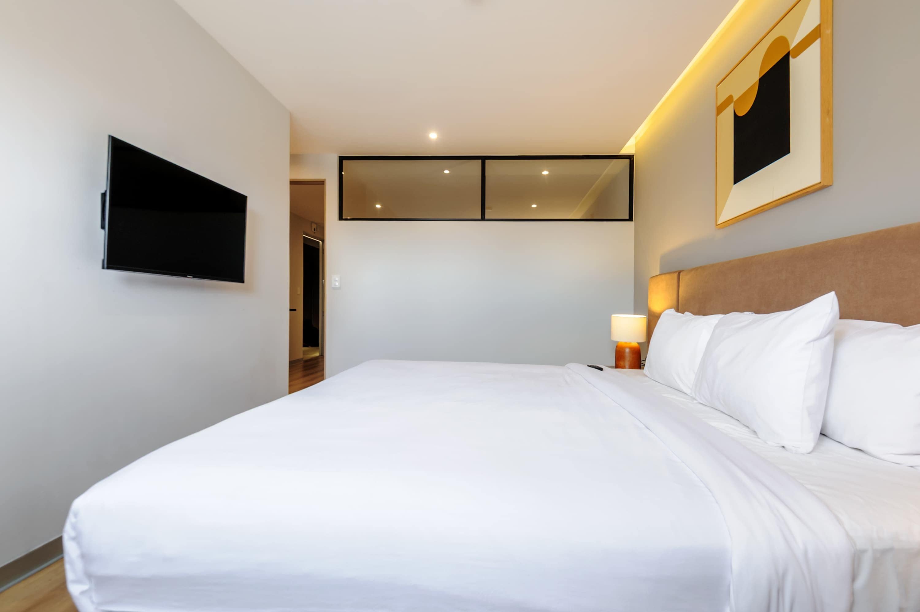 Een hotelkamer met een bed, een wandtelevisie, een nachtkastje met een lamp en een lijst boven het bed.