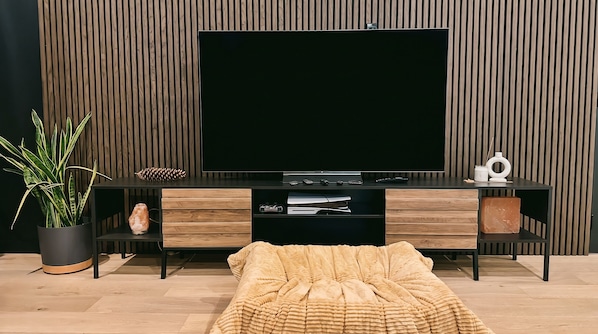 A modern living room with a large flat-screen TV, wooden furniture, a potted plant, and a cozy blanket on the floor.