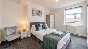 A bedroom with a bed, bedside table, chair, and artwork on the wall. - 5-Bed Flats Sleeping 11 · Free Parking · Near Beaumont Park, Huddersfield (Newsome)