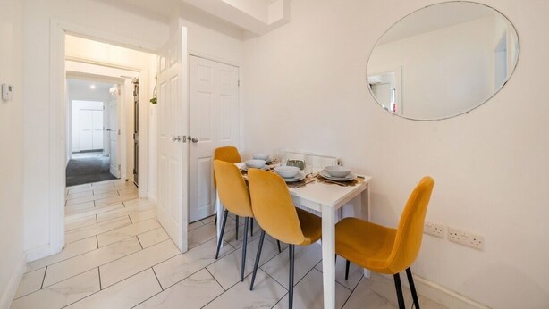 A dining area with a white table, four chairs, and a round mirror on the wall. - 5-Bed Flats Sleeping 11 · Free Parking · Near Beaumont Park, Huddersfield (Newsome)