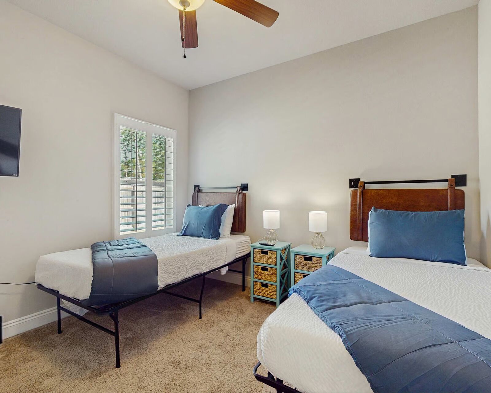 A bedroom with two beds, a ceiling fan, a window with blinds, and a small table with lamps.
