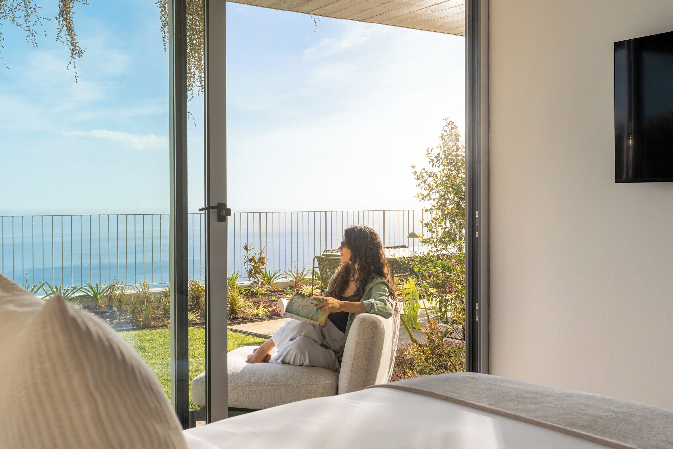 Une personne est assise sur un balcon, un livre à la main, et contemple la mer.