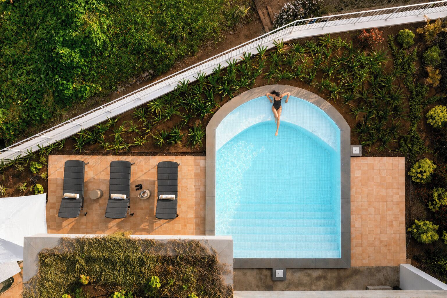 Une vue aérienne d’une piscine où l’on aperçoit une personne debout au bord, entourée de chaises longues et d’un jardin.