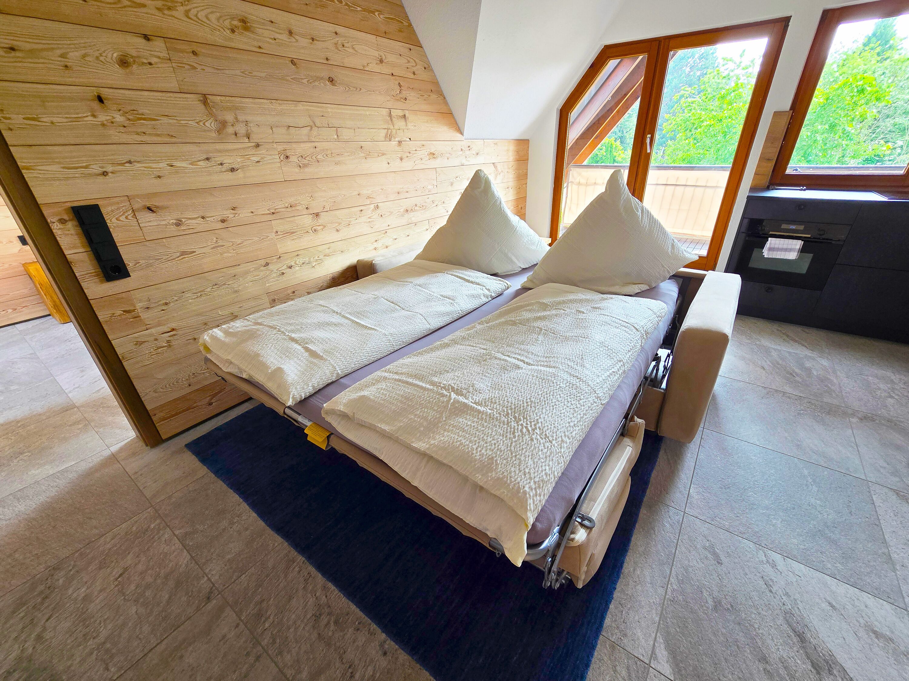 Uma cama com cabeceira de madeira, um tapete azul e vista para o exterior através de uma janela.