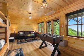 A wooden interior with a sofa, table, and foosball table. - Elevated Smoky Mountain Escape l 4,800' Views, Hot Tub & Sauna l Blue Ridge Mtn (Maggie Valley)