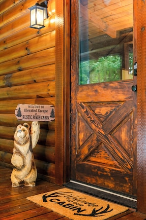 A wooden cabin entrance with a welcome sign, a bear sculpture, and a door with a geometric pattern. - Elevated Smoky Mountain Escape l 4,800' Views, Hot Tub & Sauna l Blue Ridge Mtn (Maggie Valley)