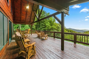 Eine Holzterrasse mit Adirondack-Stühlen und einem Tisch, mit Blick auf eine bewaldete Landschaft und Berge in der Ferne.