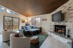 A modern living room with a fireplace, a large TV mounted on a brick wall, and a cozy seating area with a sectional sofa and armchairs. - Big Bear Cabin w/ Hot Tub, Deck & Mountain Views (Big Bear Lake)
