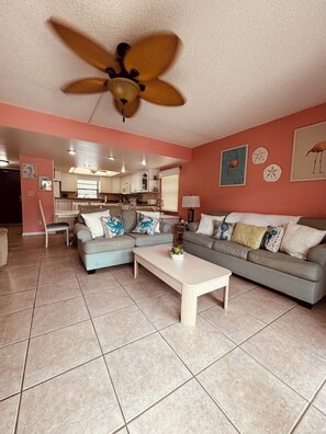 A living room with a ceiling fan, two sofas, a coffee table, and a kitchen in the background. - Beachfront 3BR Main Floor Condo/ Steps to Beach (Cocoa Beach)