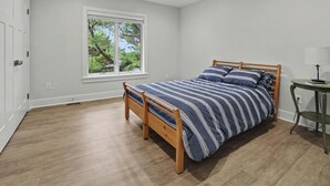 Une chambre avec un lit en bois, une fenêtre donnant sur des arbres, un ventilateur de plafond et une petite table avec une lampe.