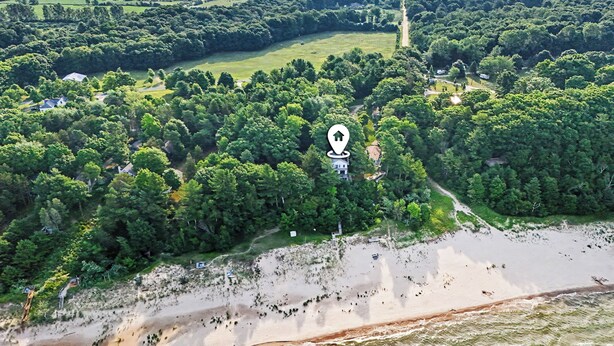 Vue aérienne d’une zone côtière avec une forêt dense, une plage de sable et un chemin dégagé menant à une maison, dont l’emplacement est indiqué par une marqueur de localisation.