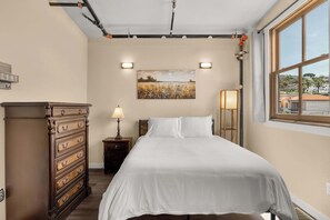 Une chambre à coucher comprenant un lit, une commode, une table de chevet, des œuvres d’art murales et une fenêtre donnant sur un paysage.