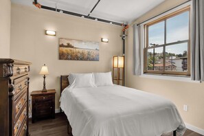 Ein Schlafzimmer mit Bett, Kommode, Nachttisch, Lampe, einem Gemälde und einem Fenster mit Blick auf Gebäude.