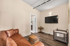 Ein Wohnzimmer mit einer Ledersofa, einer Holzkiste, einem kleinen runden Tisch mit Pflanze und einem an der Wand befestigten Fernseher.