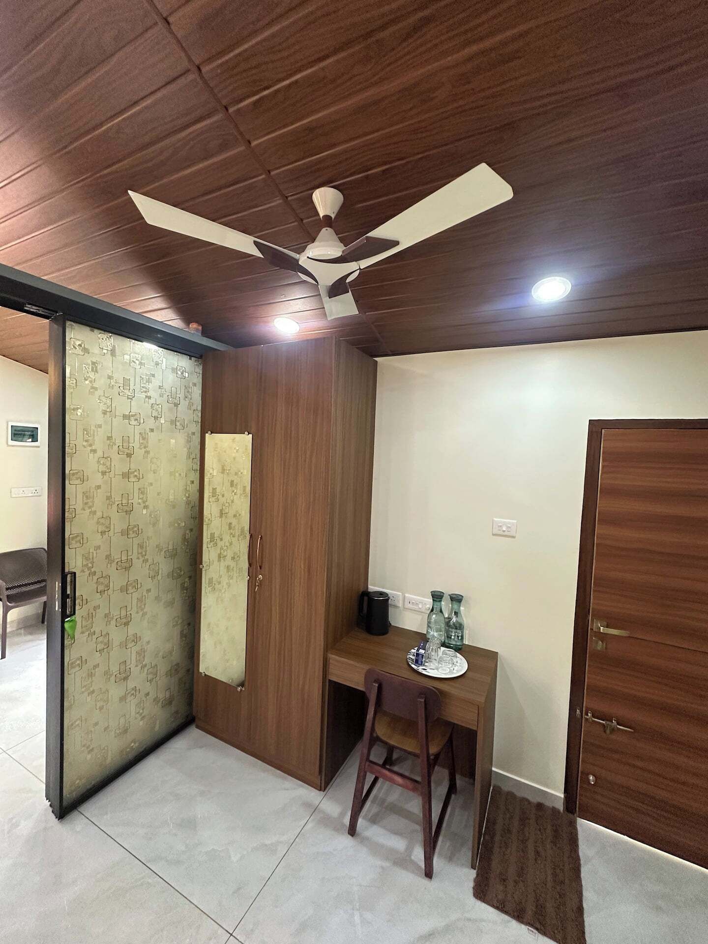 A room with a ceiling fan, a desk with a chair, a wardrobe, and a door.