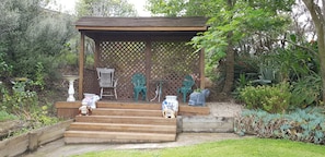 A wooden gazebo with lattice sides, surrounded by a garden with various plants and a small staircase leading up to it.
