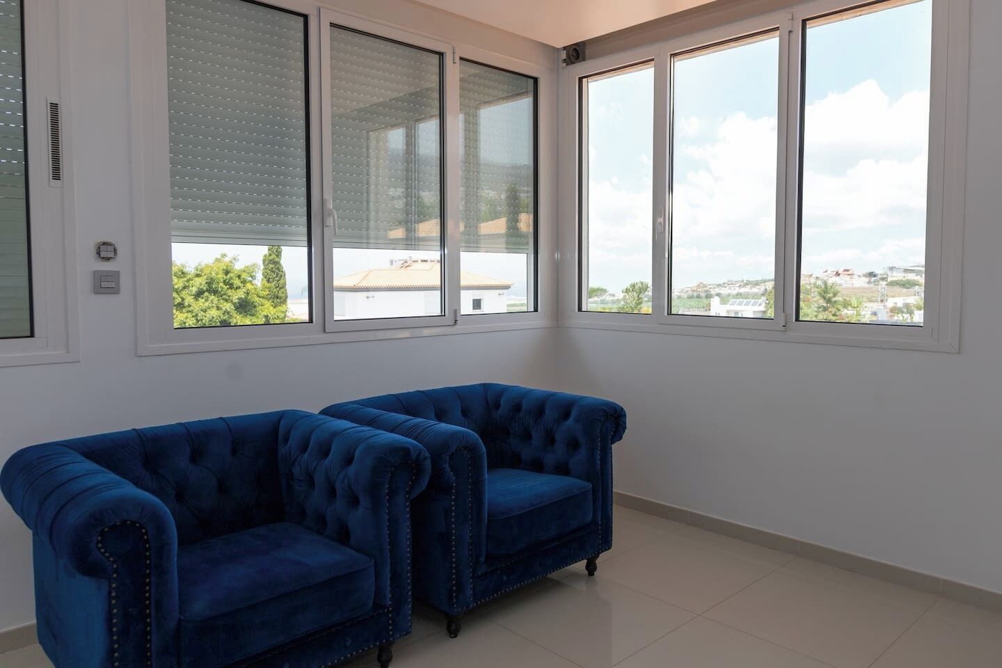 A room with two blue armchairs, a white tiled floor, and large windows with blinds.