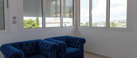 A room with two blue armchairs, a white tiled floor, and large windows with blinds.