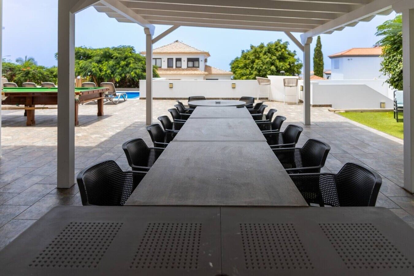 Un espace extérieur abrité, aménagé pour les repas, avec une longue table et des chaises, une piscine en arrière-plan et un bâtiment doté d’un toit en tuiles.