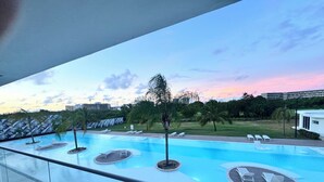A swimming pool with lounge chairs and palm trees, surrounded by a grassy area and buildings in the background. - Apartamento con balcon en Cana Bay Punta Cana (Punta Cana 23000)