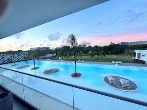 Piscina com espreguiçadeiras e palmeiras, cercada por gramado e edifícios ao fundo.