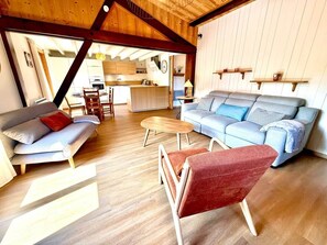 Ein gemütliches Wohnzimmer mit Sofa, Sessel und Couchtisch, Holzboden und einer Küche im Hintergrund.