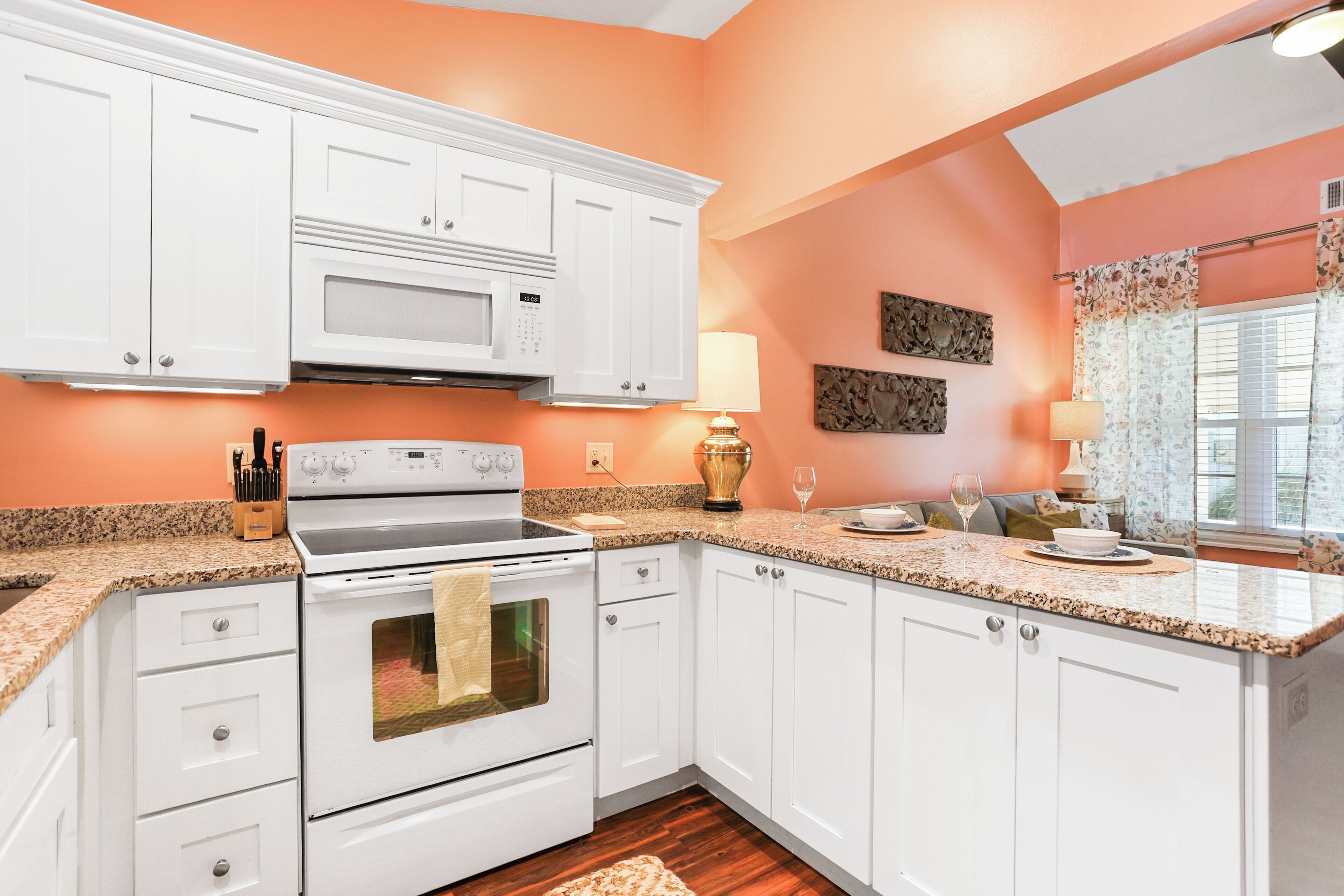 A kitchen with white cabinets, a microwave, and a stove. There is a wooden floor and a window with curtains.