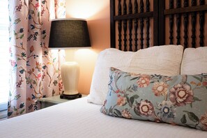 Ein Bett mit Kissen in Blumenmuster, einer Nachttischlampe und einem Vorhang mit floralem Dekor.