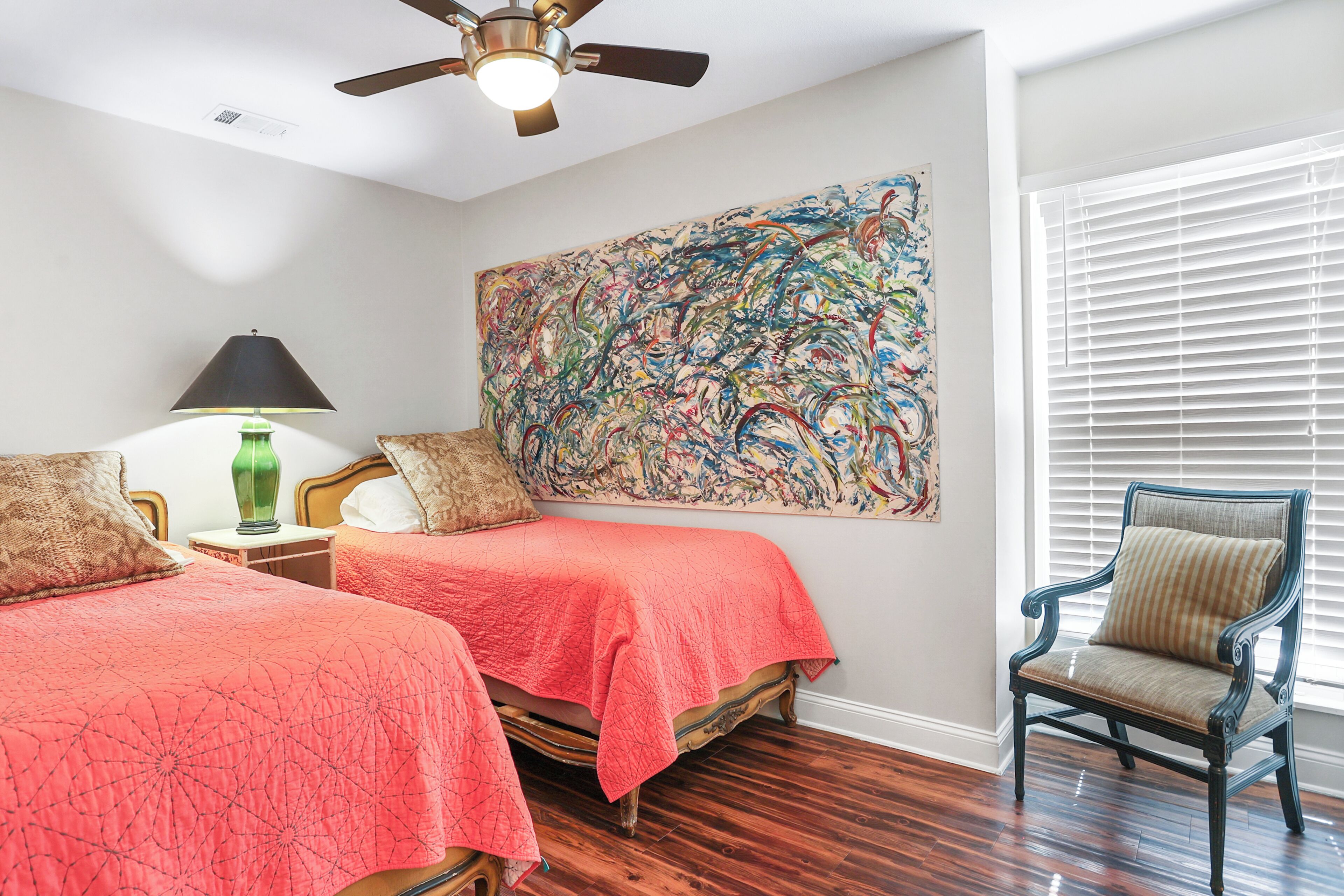 Une chambre avec deux lits, un ventilateur de plafond, un grand tableau abstrait, une chaise et une fenêtre avec des stores.